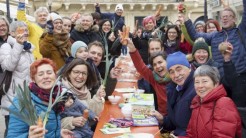 Eine Gruppe Menschen sitzt und steht um einen Tisch herum, viele halten Möhren oder Lauch hoch.